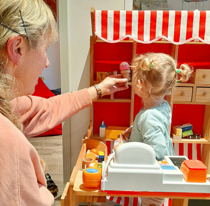 Eine Frau reicht einem kleine Kind eine Hand, das in einem Kindereinkaufsstand spielt